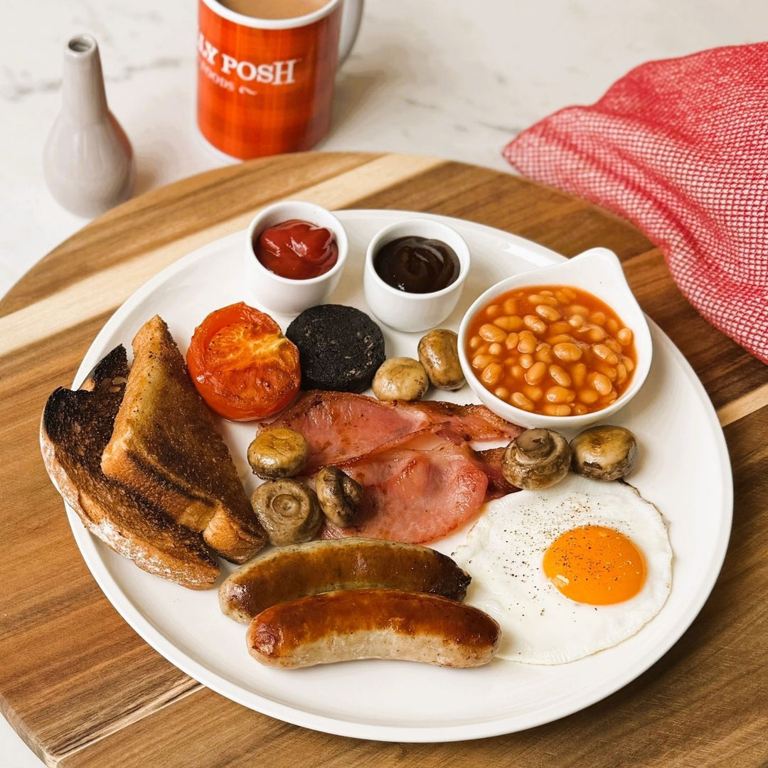 A plated full English breakfast made with Jolly Posh sausages, back bacon, black pudding, mushrooms, beans, tomato, fried egg and toast, alongside a Jolly Posh mug